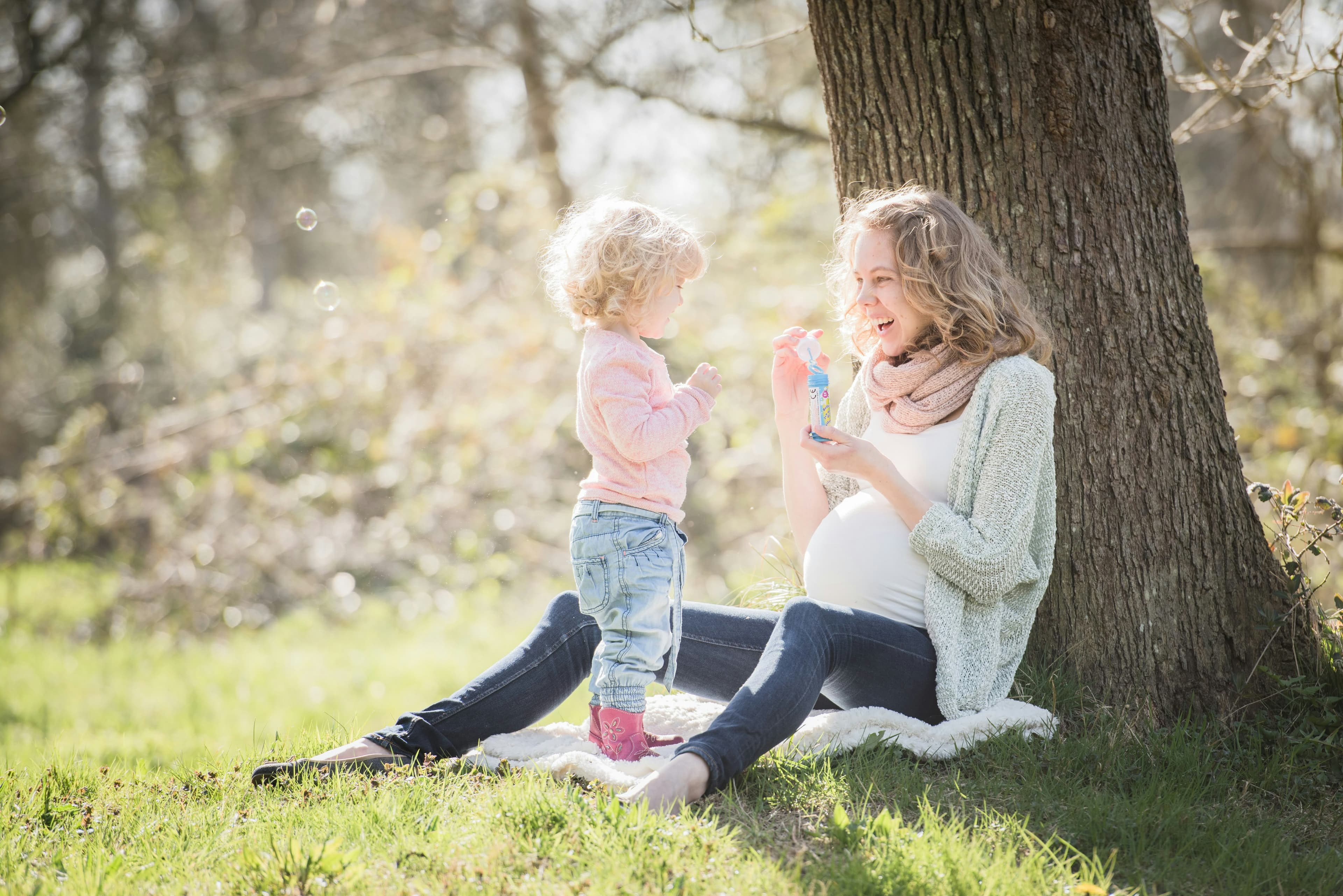 Une femme enceinte et un tout-petit sont assis sous un arbre et font des bulles de savon.